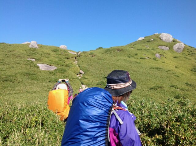晴れた日に、岩が点在する笹原の尾根を歩く登山者の後ろ姿